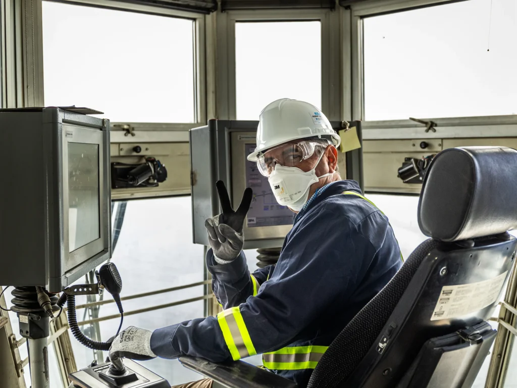 Contact St Marys Cement - ship loader operator in a hard hat and mask giving a peace sign from the loader control cab