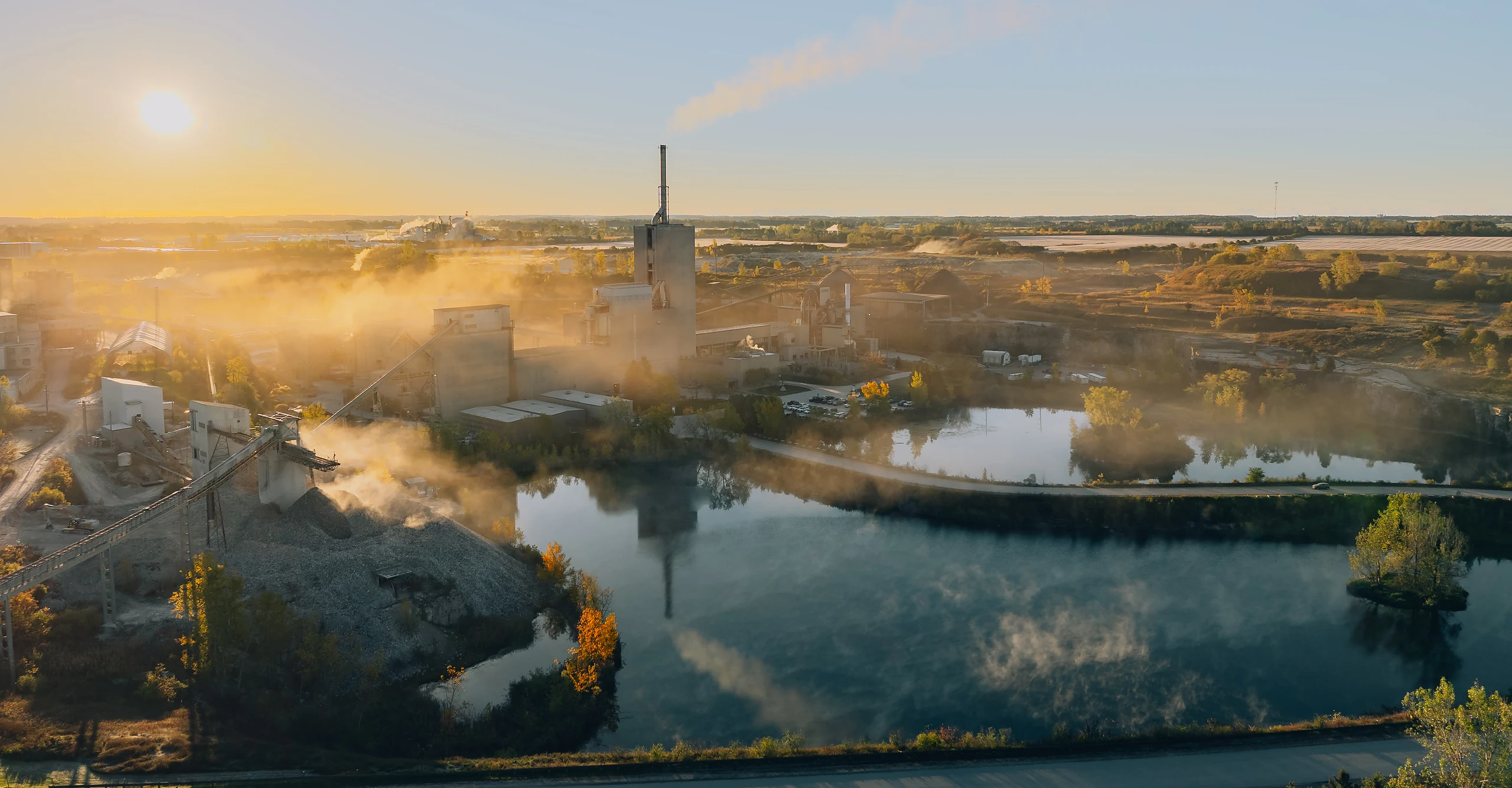 Panoramic sunrise over a quarry pond mirroring the preheater tower and its plume, mist drifting low