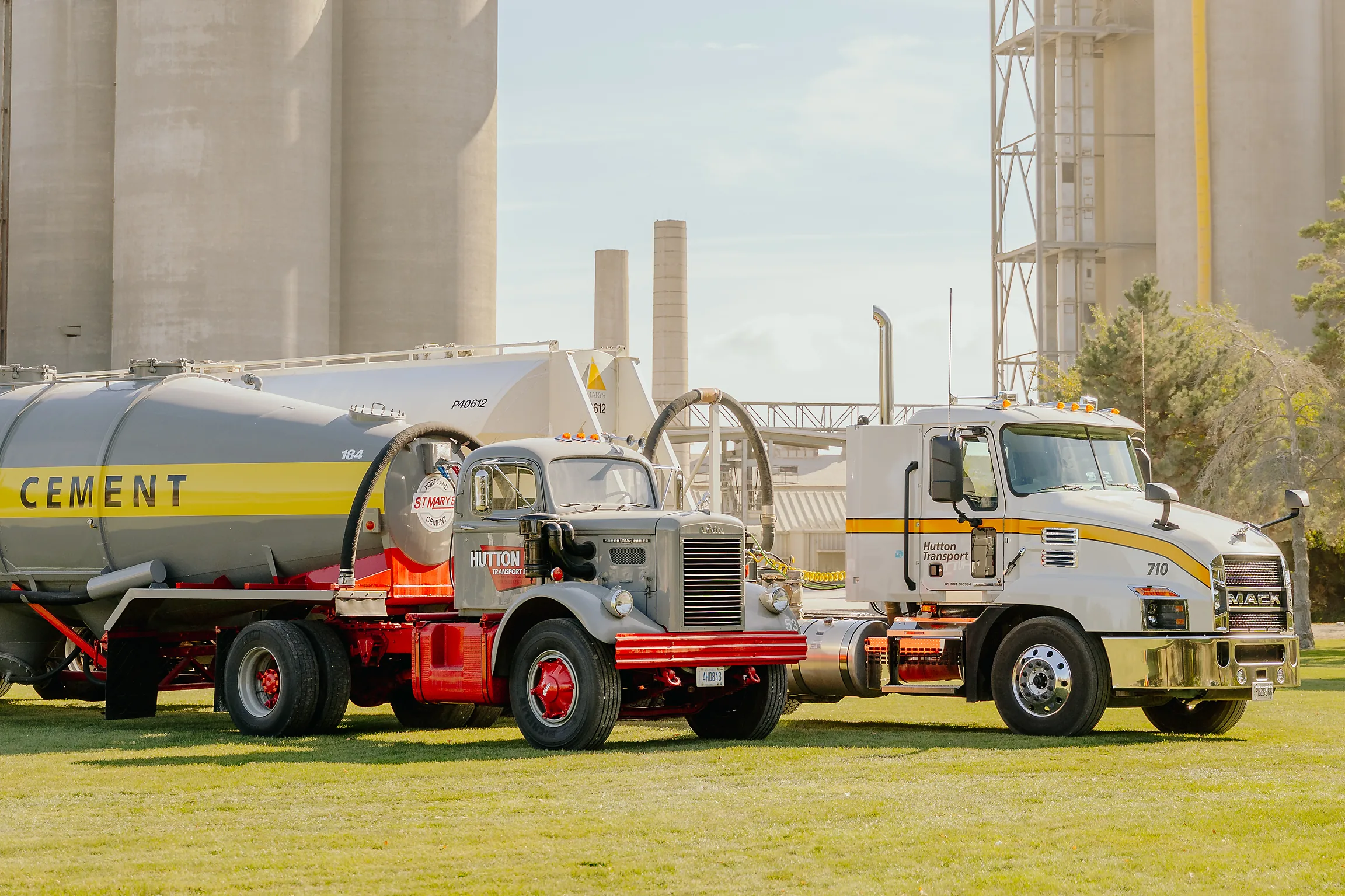 A vintage cement tanker beside a modern Hutton Transport tractor unit, with the plant silos rising behind them