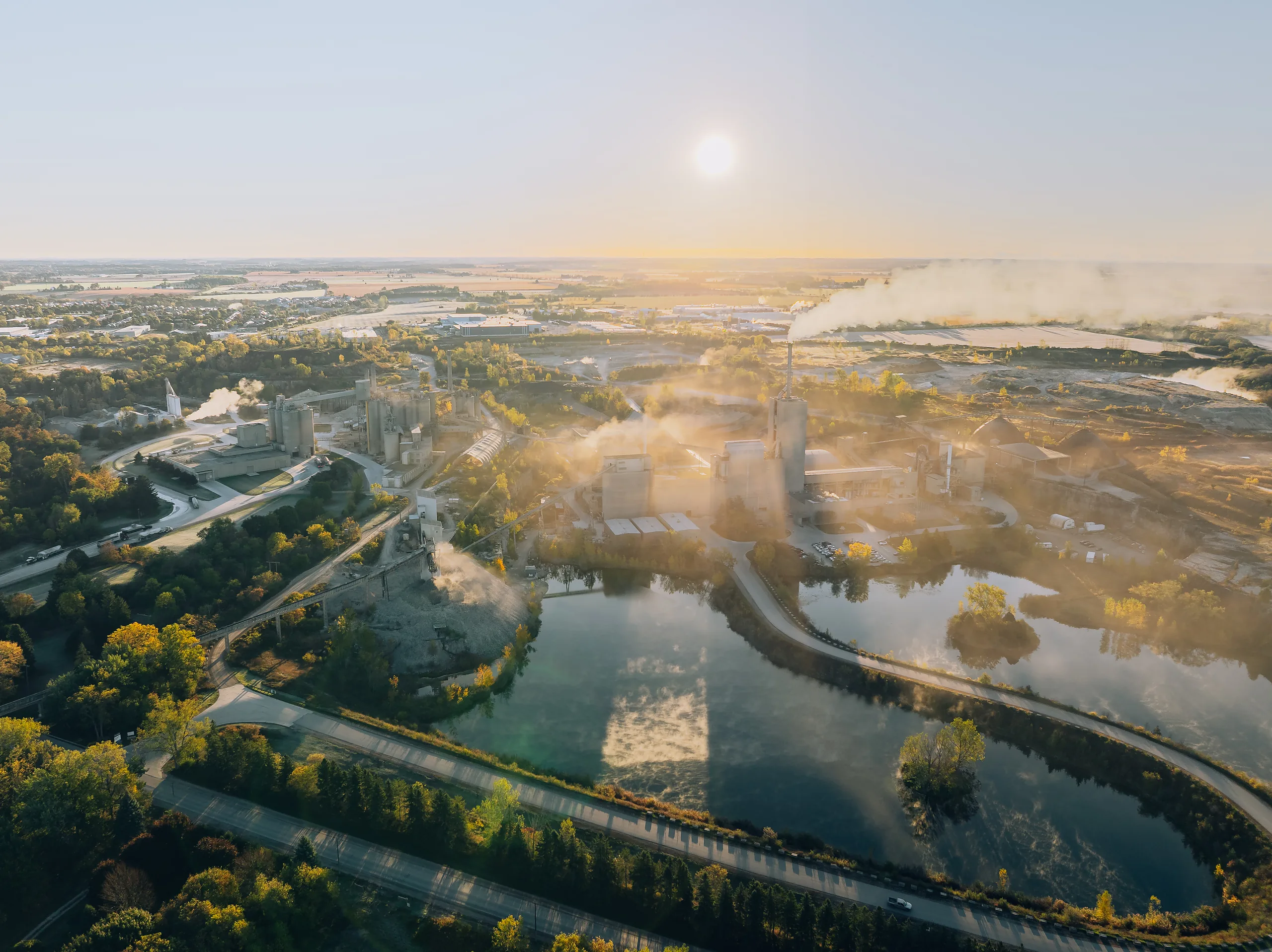 Wide morning view with silos at left, settling ponds mid-frame and a tree-lined highway in the foreground