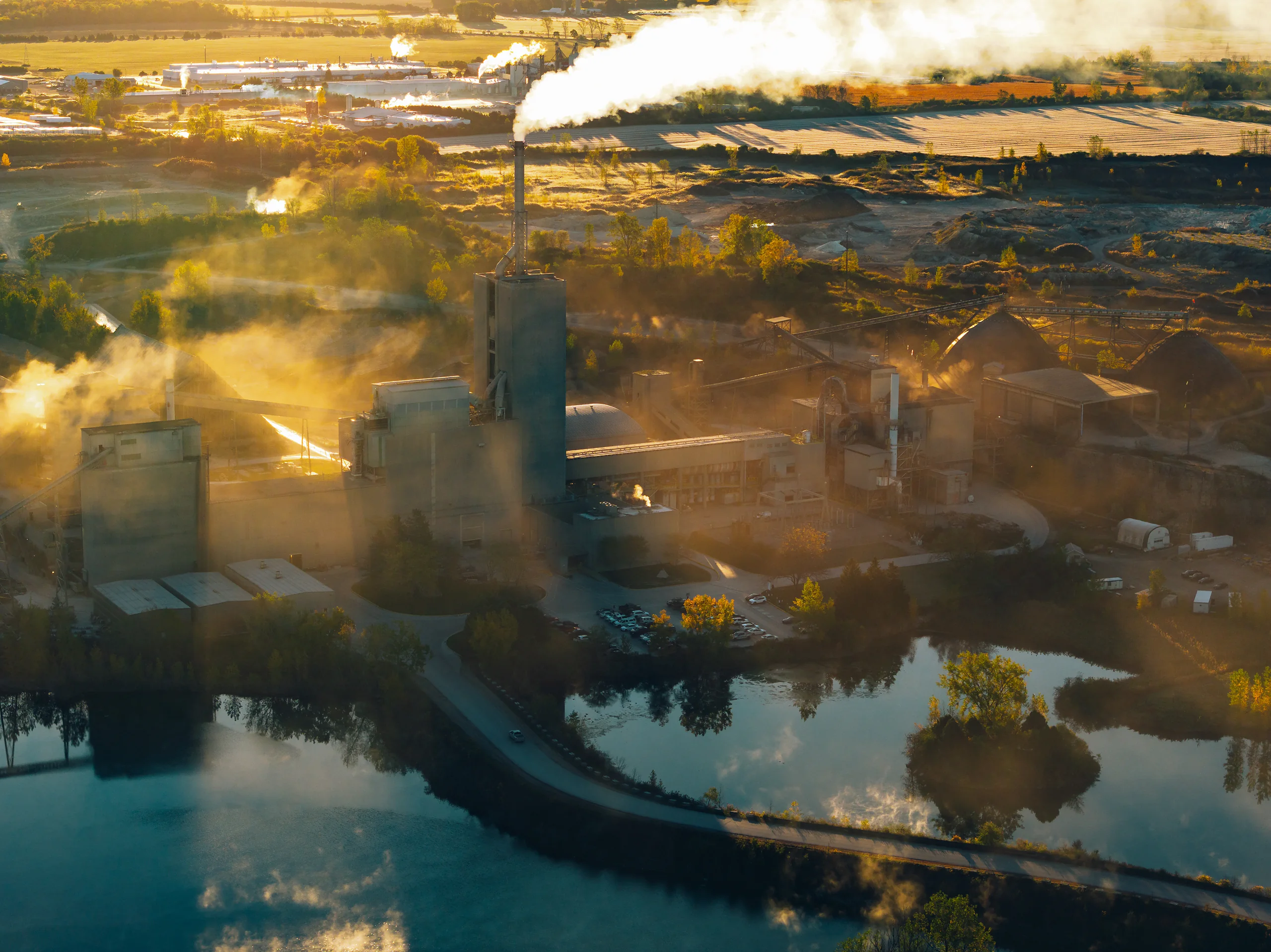 Backlit view of the kiln stack trailing a long plume, with a turquoise quarry pond in the lower foreground